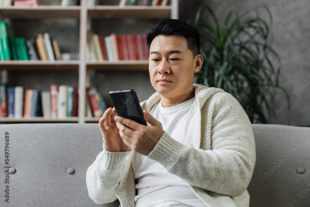 Attractive asian man in casual wear sitting on comfy couch with modern smartphone in hands. Smiling male person surfing internet during leisure time at home.