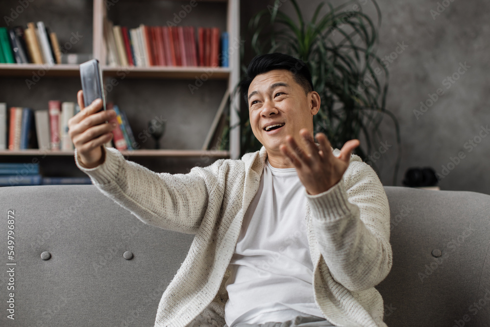 Attractive asian man in casual wear sitting on comfy couch with modern smartphone in hands. Smiling male person having video call during leisure time at home.