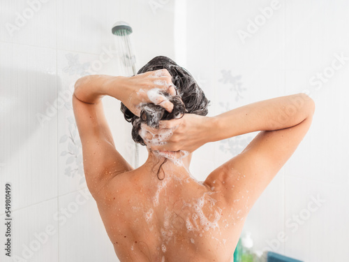 Naked woman with long hair takes a shower. Woman washes her hair with shampoo. Morning routine in modern urban apartment. White bathroom.