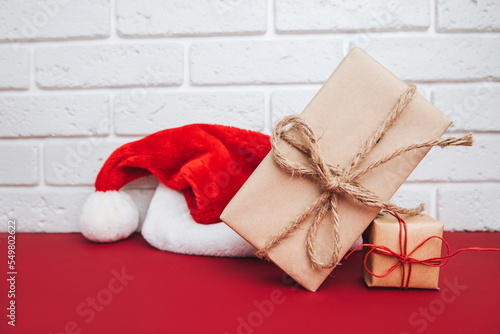 Gifts in brown packaging on a red table with Christmas cap on background. Merry Christmas background. Front view