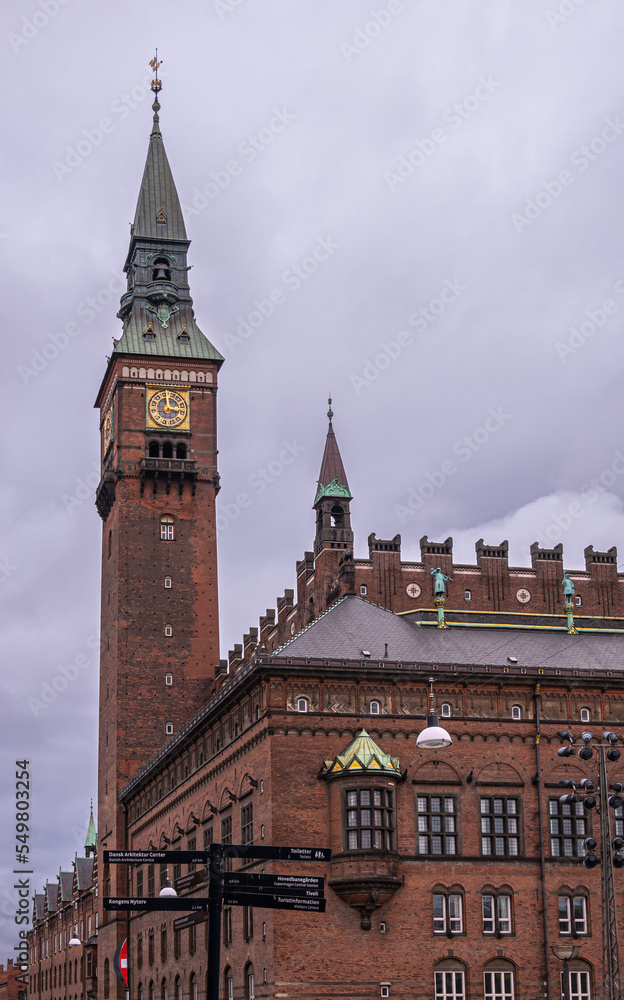Copenhagen, Denmark - July 23, 2022: NW corner of red brick town hall with its tall bell tower, with golden clock and sharp green spire, against gray cloudscape on Radhuspladsen