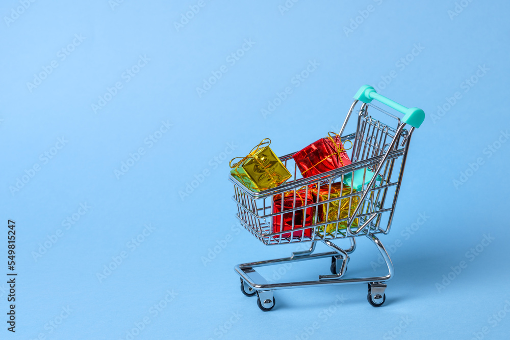 Close up of a small shopping cart full of colourful Christmas gifts on blue background. Copy space. Close up of a small shopping cart full of colourful Christmas gifts on blue background. Copy space.