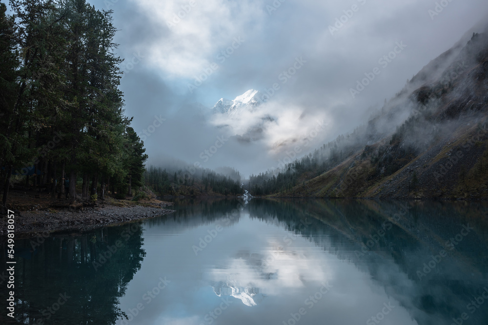 Fototapeta premium Tranquil scenery with snow castle in clouds. Mountain creek flows from forest hills into glacial lake. Snowy mountains in fog clearance, small river and coniferous trees reflected in calm alpine lake.