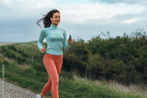 An attractive young woman is running in nature wearing fitness clothes and listening to music on her smartphone.