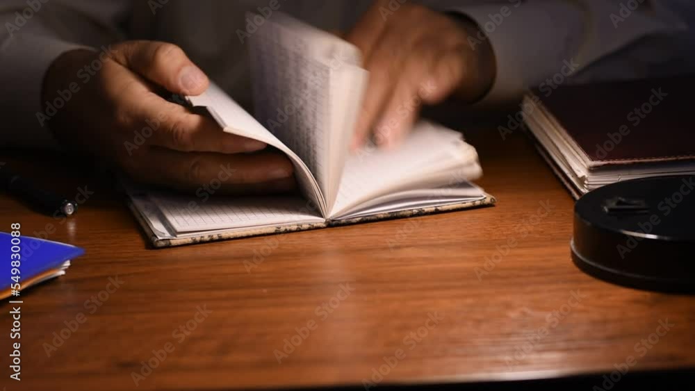 The man is sitting at the table. He opens the notepad, finds the right page and starts writing in it. The man is sitting at the table. He opens the notepad, finds the right page and starts writing in it.
