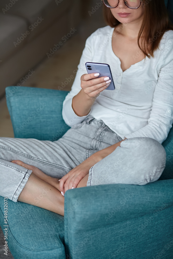 Young woman using mobile phone holding in hands while sitting in armchair at home Young woman using mobile phone holding in hands while sitting in armchair at home