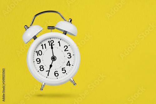 A white vintage alarm clock shows 7 o'clock, casting a shadow on a yellow background. The concept of morning and early awakening. Time to act or play sports. Place with copy space.