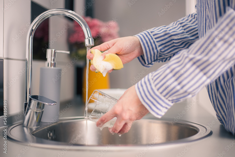 Woman washing glass with sponge over sink, closeup. Maid using a liquid detergent with a sponge to wash drinking glasses and dishes