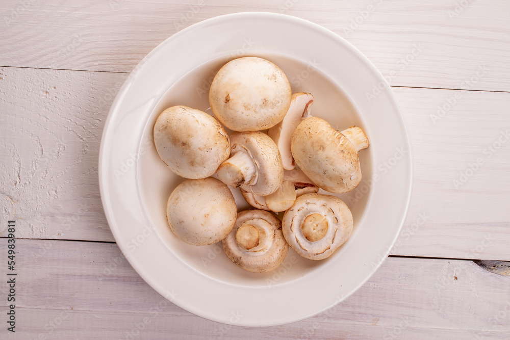Several organic fresh appetizing mushrooms champignon in a white ceramic plate, close-up, on a table of natural wood, painted white, top view. Several organic fresh appetizing mushrooms champignon in a white ceramic plate, close-up, on a table of natural wood, painted white, top view.