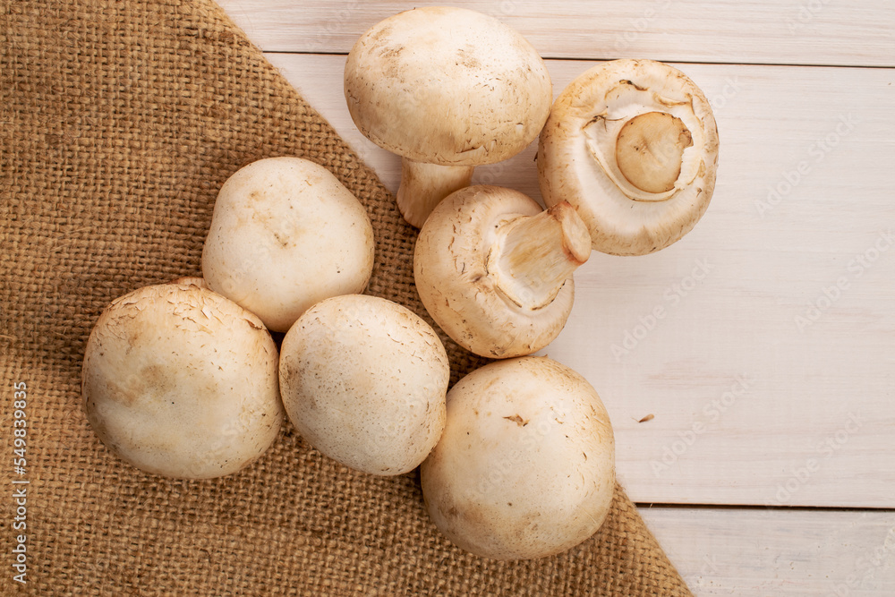 Several whole organic fresh appetizing mushrooms champignon with a jute napkin, on a table made of white painted natural wood, top view.