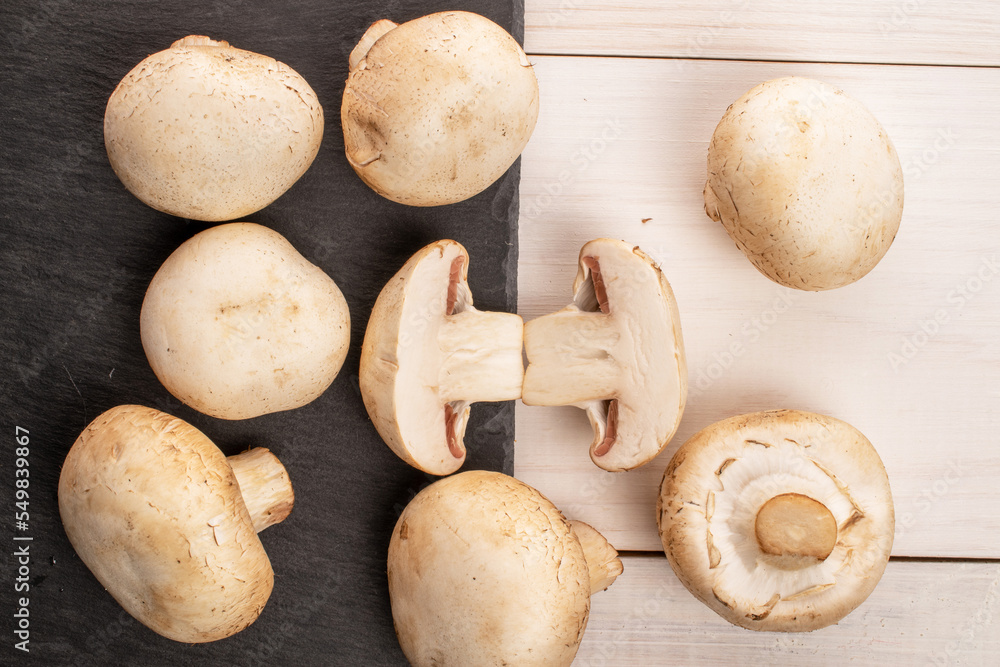 A few whole and two halves of organic fresh appetizing mushrooms champignon with a serving board of slate on a table made of white painted natural wood, top view. A few whole and two halves of organic fresh appetizing mushrooms champignon with a serving board of slate on a table made of white painted natural wood, top view.
