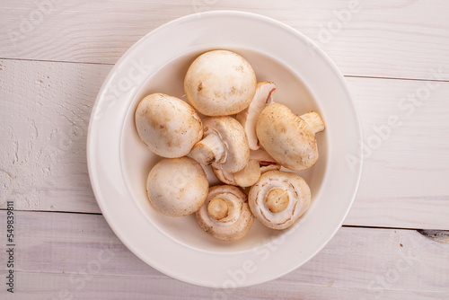 Several organic fresh appetizing mushrooms champignon in a white ceramic plate, close-up, on a table of natural wood, painted white, top view.