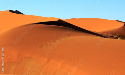 Fototapeta Naklejka Na Ścianę i Meble -  Close up of a bright orange sand dune showing the ripples in the sand caused by the wind, with a natural pale blue sky