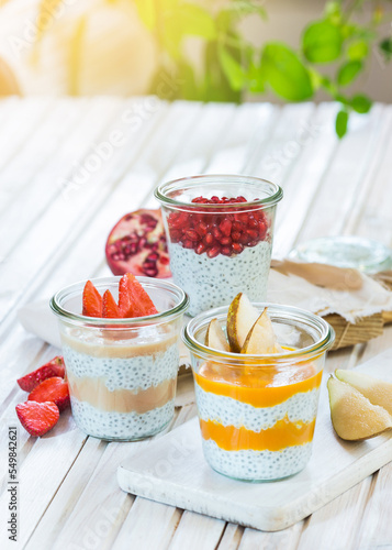 Breakfast chia desserts in jars with fresh fruit - pomegranate, pear and strawberry on a white, wooden board