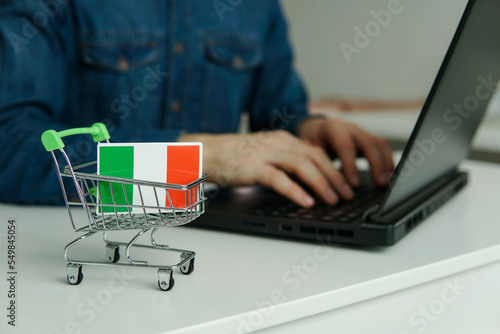 Small shopping cart with flag of Italy on the table. Man using laptop for shopping online.