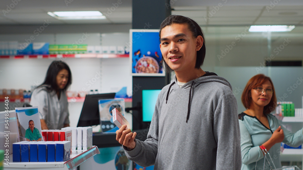 Asian man reading leaflet on pills box in pharmacy, checking packages of drugs and pharmaceutics. Young client visiting medical retail store to buy medicaments and vitamins. Handheld shot.
