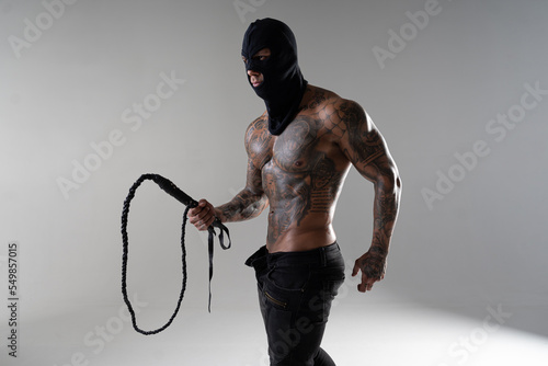 Muscular shirtless young man with whip in a mask.Brutal handsome man with tattooed body. Men tattoo casual fashion. Portrait of handsome male model. Muscular athletic sexy male with naked torso.