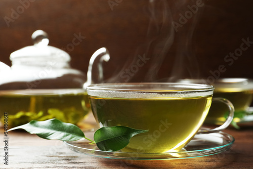 Fresh green tea in glass cup with saucer and leaves on wooden table, closeup