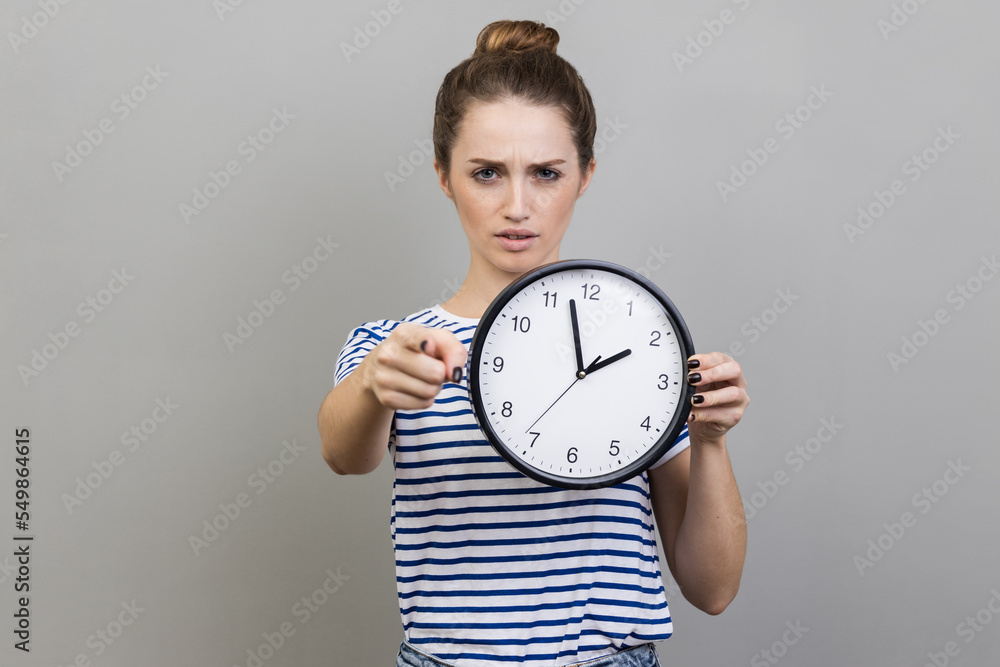Portrait of bossy serious woman wearing striped T-shirt pointing finger at you holding in hand big wall clock, motivation. Indoor studio shot isolated on gray background. Portrait of bossy serious woman wearing striped T-shirt pointing finger at you holding in hand big wall clock, motivation. Indoor studio shot isolated on gray background.