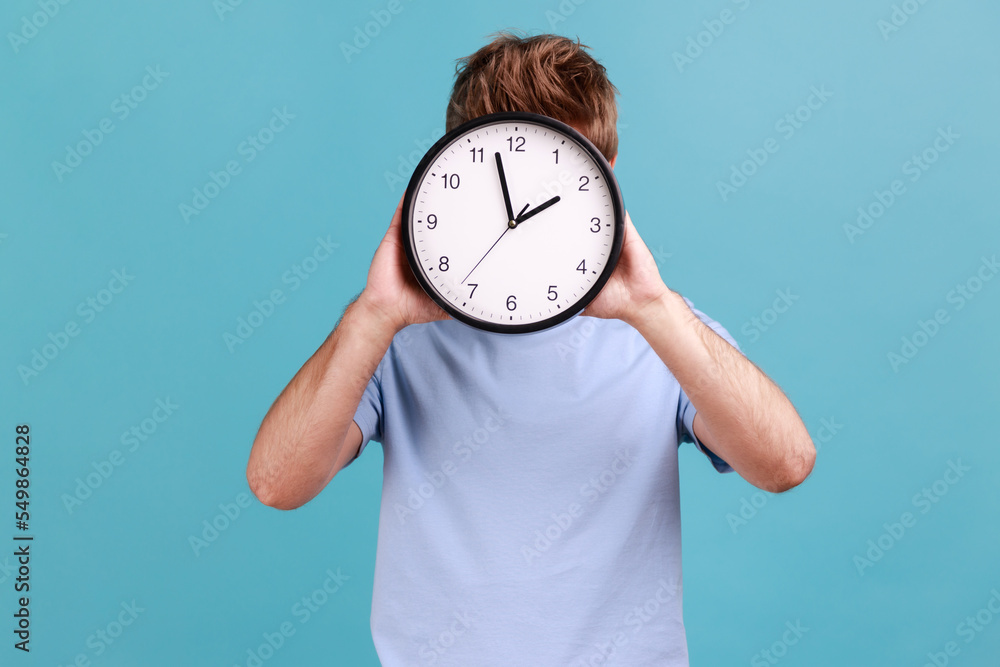 Portrait of unknown anonymous man holding big wall clock hiding his face, time management, schedule and meeting appointment. Indoor studio shot isolated on blue background. Portrait of unknown anonymous man holding big wall clock hiding his face, time management, schedule and meeting appointment. Indoor studio shot isolated on blue background.