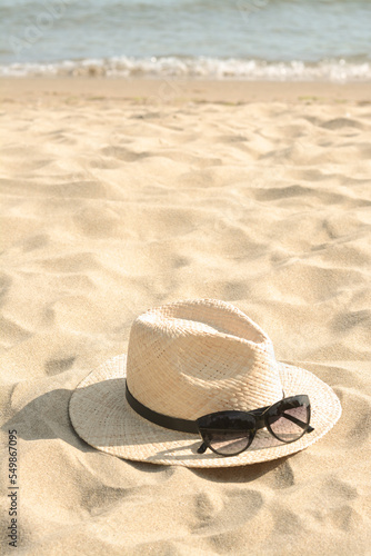 Hat with beautiful sunglasses on sand near sea. Space for text