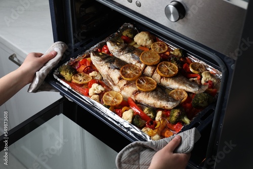 Woman taking out baking tray with sea bass fish and vegetables from oven, closeup