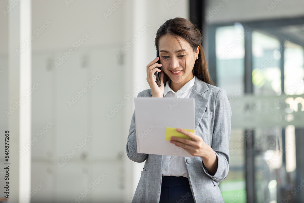 Young smiling Business asian woman talking on mobile phone, standing in workplace office with document in her hand Young smiling Business asian woman talking on mobile phone, standing in workplace office with document in her hand