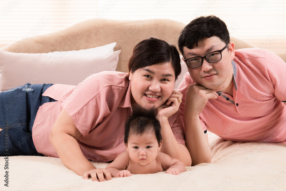 Fototapeta premium Happy asian family laying down and taking pictures together in bedroom. parents and little daughter looking at camera posing for photo. Exemplary family portrait, love and bond concept