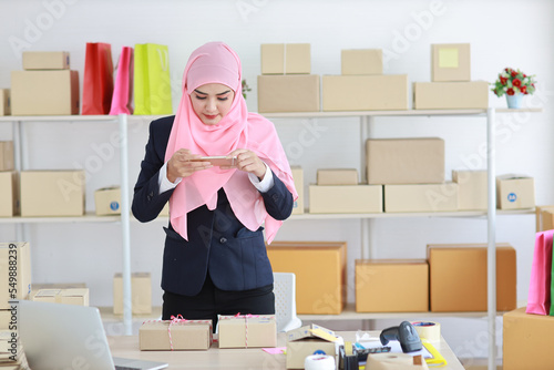 eligious asian woman in blue muslim suit standing and taking picture of package box deliver from mobile phone. Startup small business SME freelance girl work at home with happy smiling face photo