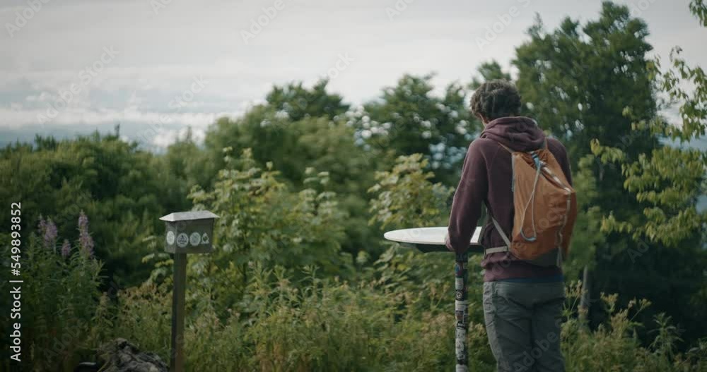 A young hiker with an orange backpack is standing by a signpost reading and looking in different directions. At the end hiker leaves the singpost and continues his jurney. Cloudy summer day.