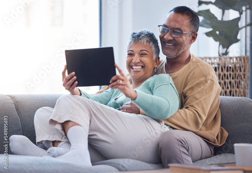 Tablet, love and relax with a senior couple sitting on a sofa in the living room of their home together. Funny, joke and humor with a mature man and woman laughing while watching a comedy in a house