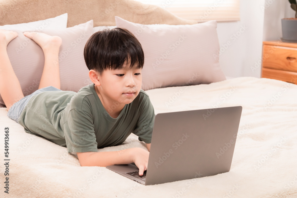Selective focus Asian little child boy laying down on bed using laptop playing games, watching movie, favorite manga or searching internet with curious alone happy at home, natural light Selective focus Asian little child boy laying down on bed using laptop playing games, watching movie, favorite manga or searching internet with curious alone happy at home, natural light