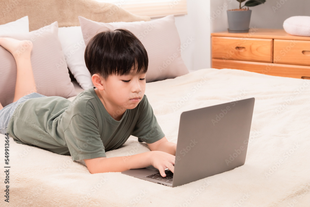 Selective focus Asian little child boy laying down on bed using laptop playing games, watching movie, favorite manga or searching internet with curious alone happy at home, natural light Selective focus Asian little child boy laying down on bed using laptop playing games, watching movie, favorite manga or searching internet with curious alone happy at home, natural light