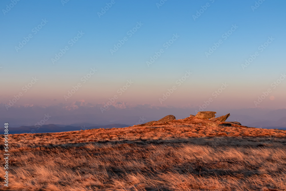 Scenic view of rock formation and Karawanks mountains at sunrise seen from alpine meadow at Ladinger Spitz, Saualpe, Lavanttal Alps, Carinthia, Austria, Europe. Early morning golden hour in Wolfsberg