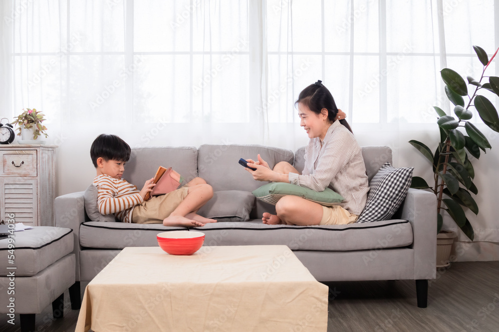 Asian single mother sitting on sofa relax using smartphone shopping online while handsome son laying down play game with tablet next to mom. Family using different electronic devices. enjoy social. Asian single mother sitting on sofa relax using smartphone shopping online while handsome son laying down play game with tablet next to mom. Family using different electronic devices. enjoy social.