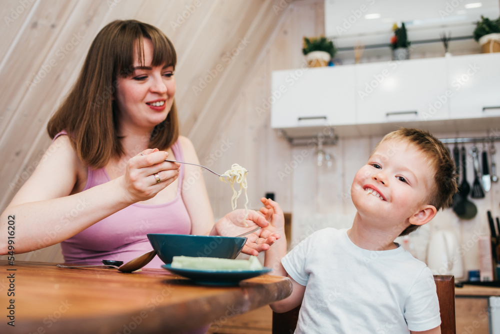 Cheerful child eating pasta in the kitchen with mom Cheerful child eating pasta in the kitchen with mom