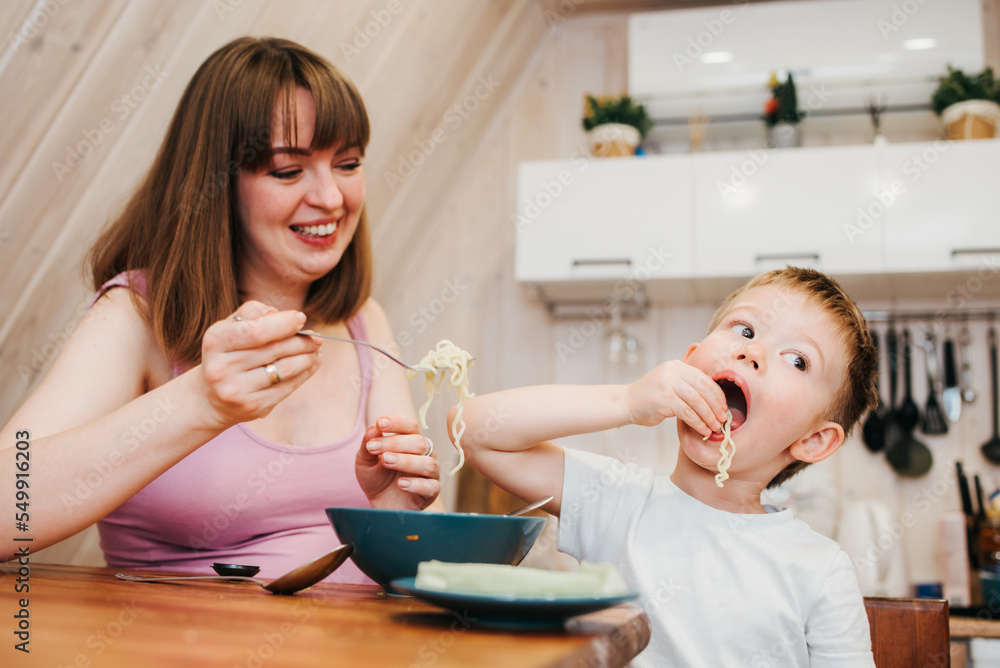 Cheerful child eating pasta in the kitchen with mom