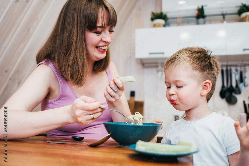 Cheerful child eating pasta in the kitchen with mom Cheerful child eating pasta in the kitchen with mom