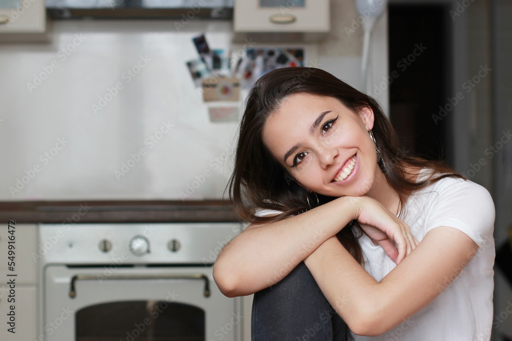 A beautiful young brunette girl looks directly into the camera and smiles with a wide snow-white smile. The kitchen is visible in the background