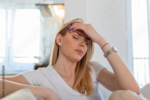 Portrait of a young blond woman sitting on the couch at home with a headache and migraine . Beautiful woman suffering from chronic daily headaches. Sad woman holding her head because sinus pain