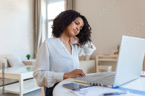 Portrait of young stressed woman sitting at home office desk in front of laptop, touching aching back with pained expression, suffering from backache after working on pc