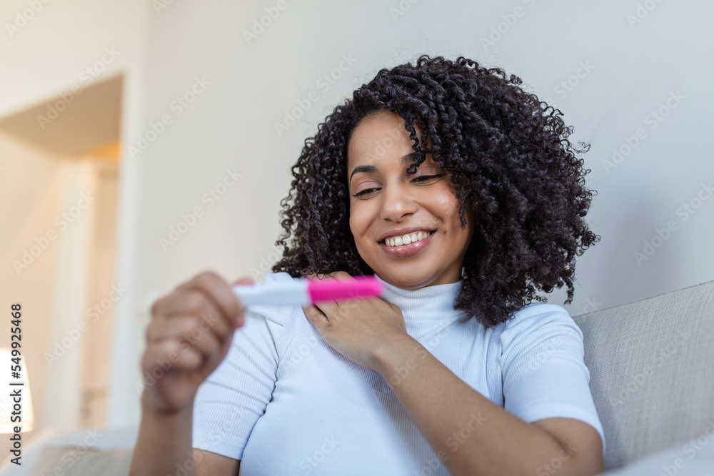 Young woman looking at pregnance test in happiness. Finally pregnant. Attractive black women looking at pregnancy test and smiling while sitting on the sofa at home Young woman looking at pregnance test in happiness. Finally pregnant. Attractive black women looking at pregnancy test and smiling while sitting on the sofa at home