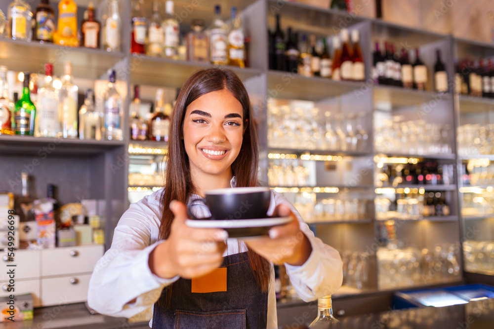 Beautiful female barista is holding a cup with hot coffee, looking at camera and smiling while standing near the bar counter in cafe