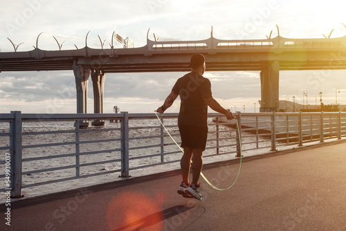 Tela Rear view athlete man working out on skipping rope in urban embankment outdoors at sunset background