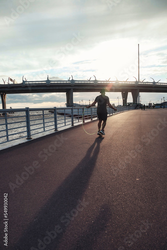 Rear view sportsman workout on skipping rope in urban embankment outdoors at sunset background. Athletic man working out with jumping rope in city district. Healthy lifestyle concept. Copy text space