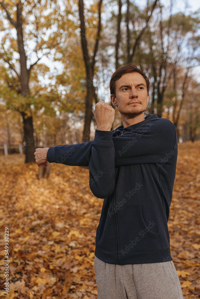 Confident sportsman stretching arms and warming up while preparing for training in autumn park Confident sportsman stretching arms and warming up while preparing for training in autumn park