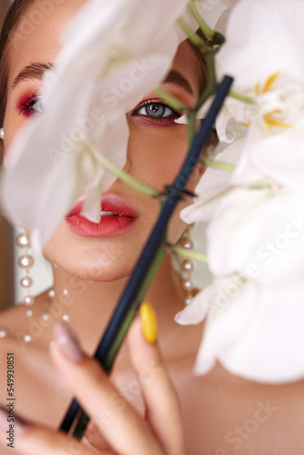 Headshot of pretty woman with pearls on face hiding behind white orchid flowers and looking at camera 