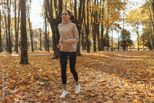 Full length of motivated young woman running along path in park while enjoying training on sunny day in autumn 