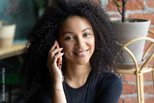Black happy woman with African curls speaking on cellphone at home or cafe indoors. Communication with friends of relatives. Comfort relationships. Freelance