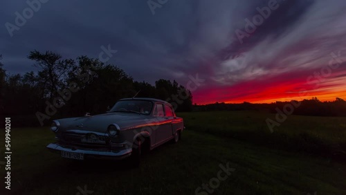 Old historic Gaz 21 car standing on a grassy clearing. In the background, an amazing sunrise in a time lapse shot with a red glow and clouds floating across the sky photo